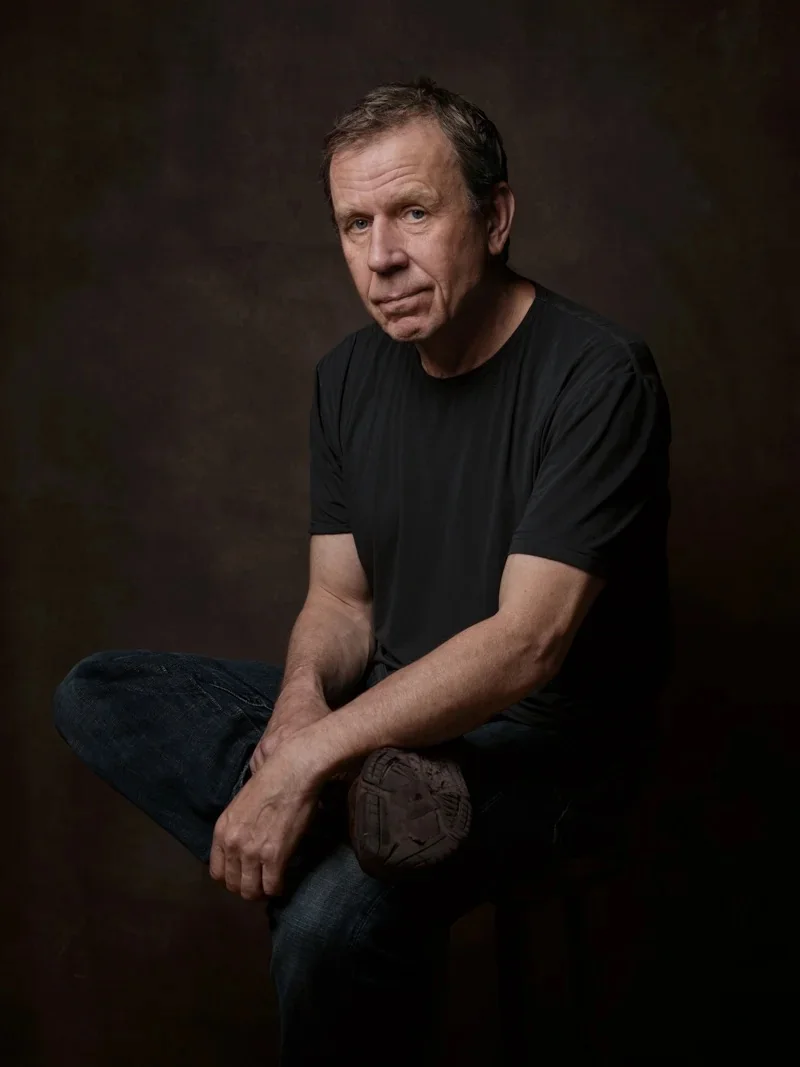 Full-body portrait of a man sitting cross-legged on a stool against a dark brown background.